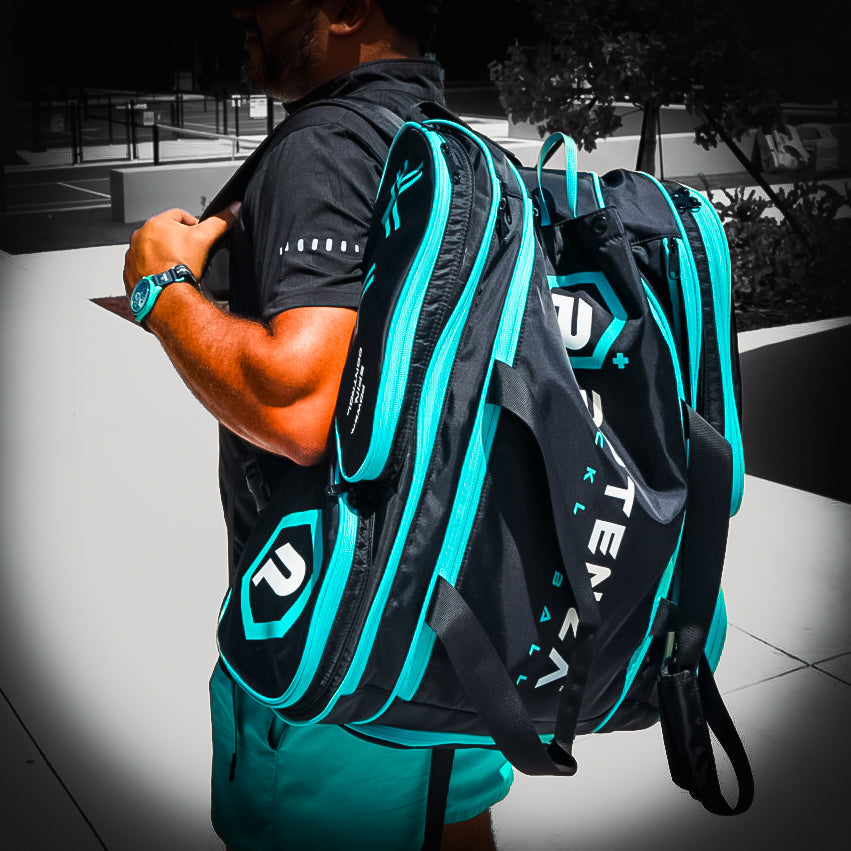 A man outdoors in bright sunlight near a tennis court carries two Potenza Pickleball Tournament Paddle Bags with paddle protection and bold “P” logos, plus “TEAM” text on his back, holding one strap in his right hand.