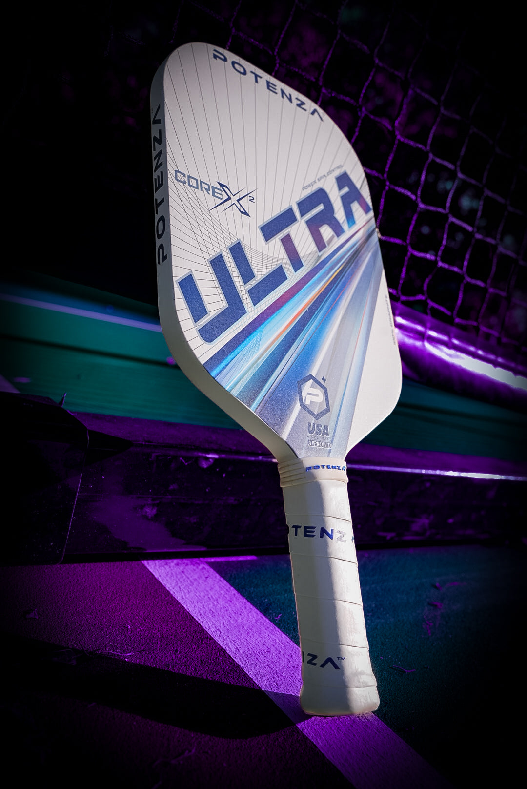 A white Potenza Pickleball ULTRA+ COREx2 paddle with a textured handle and USA marking leans against the net under dramatic purple lighting, with the court’s edge and net visible.