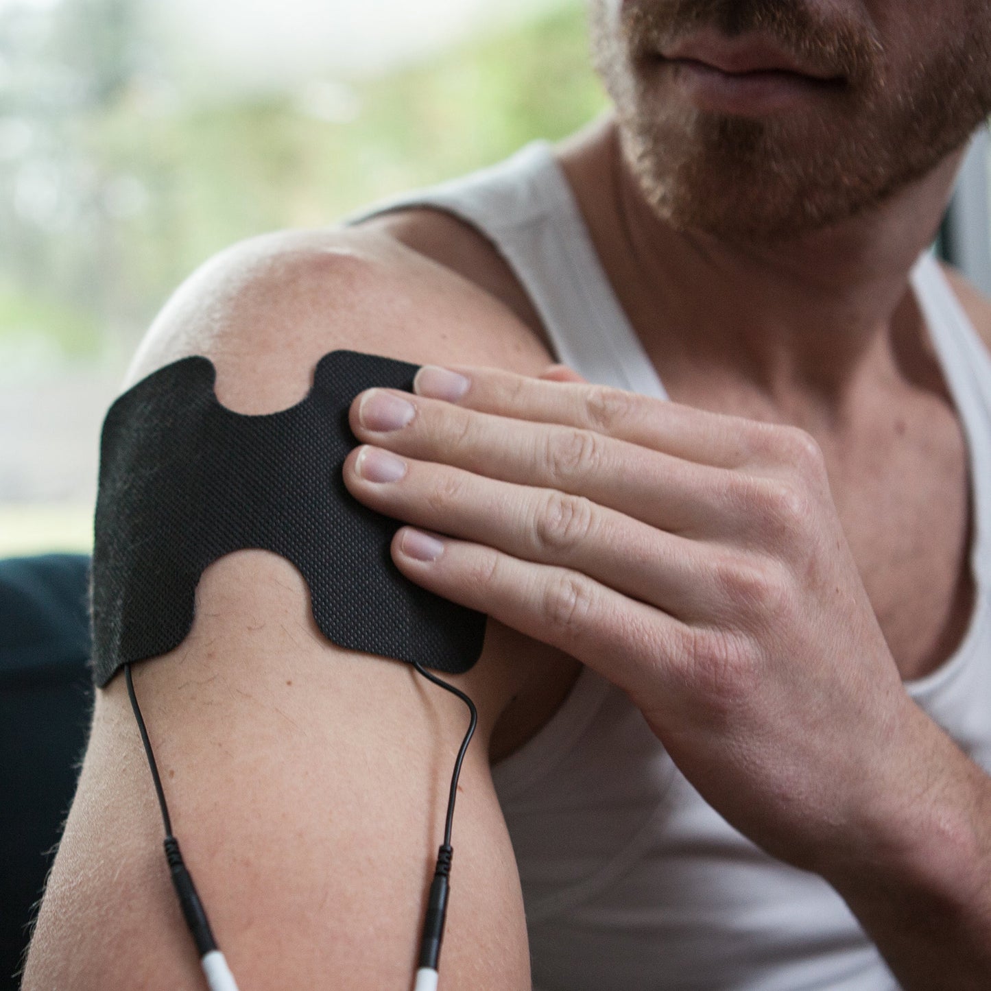 A man in a white tank top uses the iReliev Wired Large & Small Refill Kit, attaching a large black electrode pad with wires to his upper arm for electrotherapy or muscle stimulation. His face is partially visible indoors near a window.