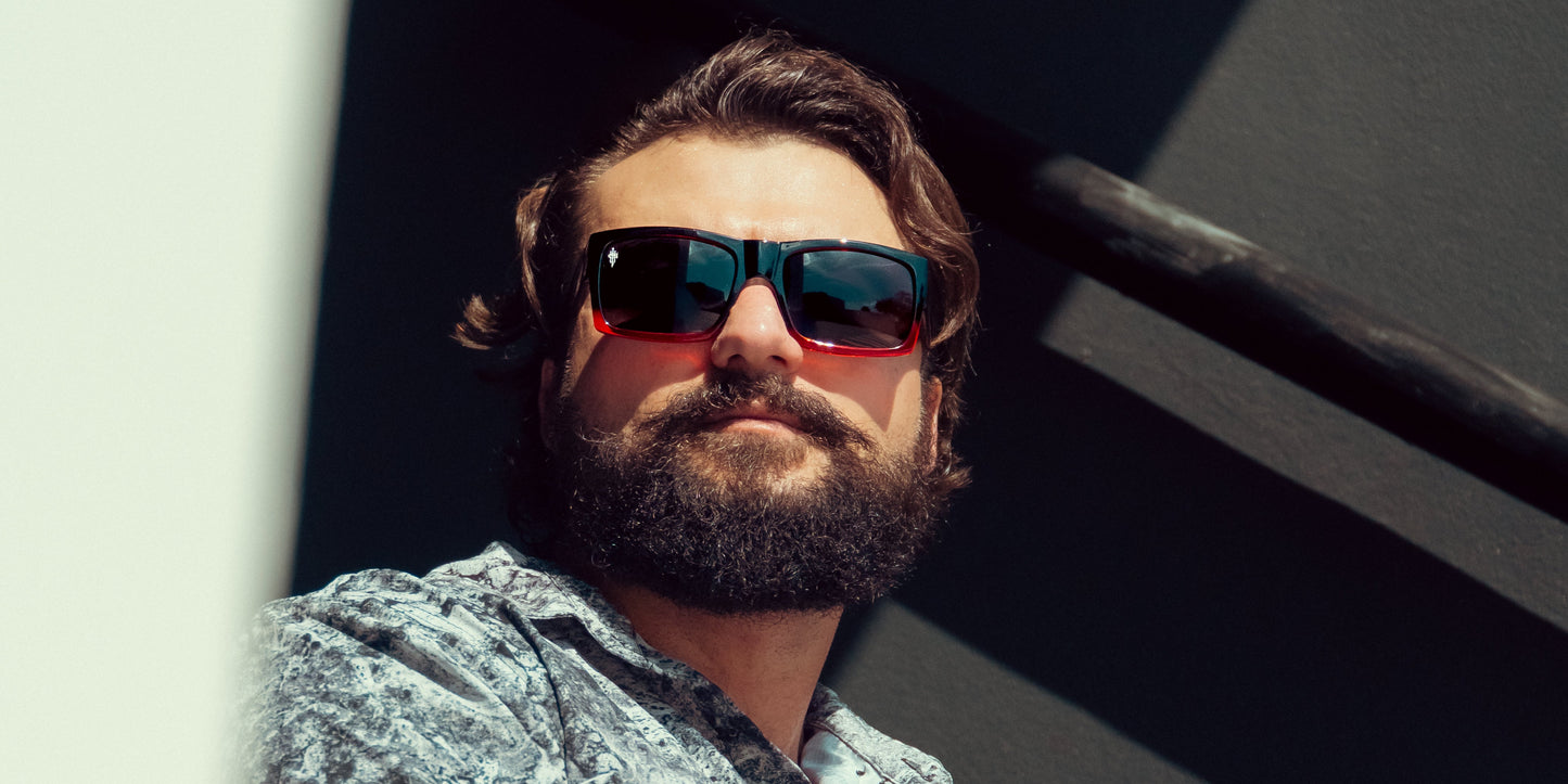 A bearded man with wavy brown hair wears Neven Eyewear’s Blood Fade sunglasses—large, dark, and stylish—with a patterned shirt. He looks up as sunlight casts shadows, framed by a dark wall and railing in the background.