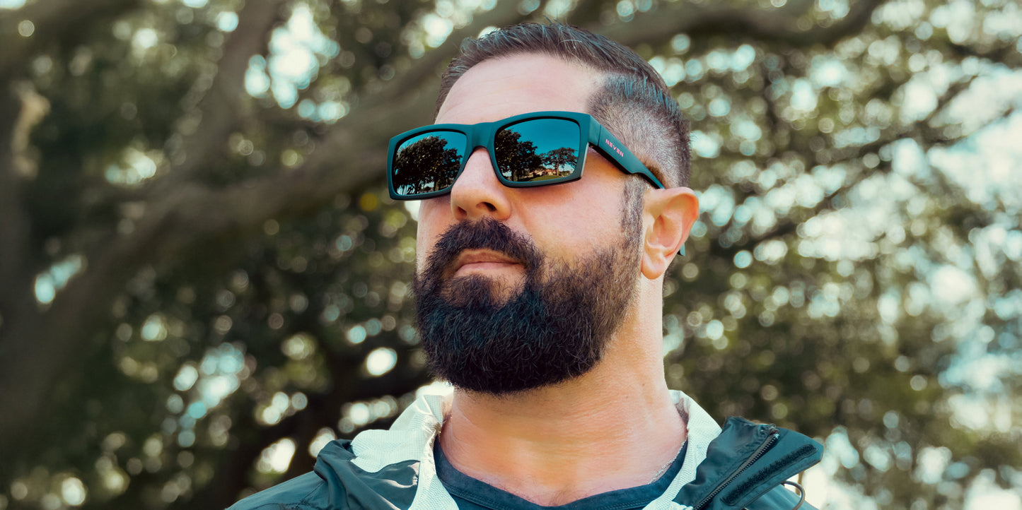 A bearded man with short hair wears Neven Eyewear Redrum sunglasses and a dark jacket outdoors. Sunlight filters through green trees, reflecting in the polarized matte black lenses as he confidently gazes upward.