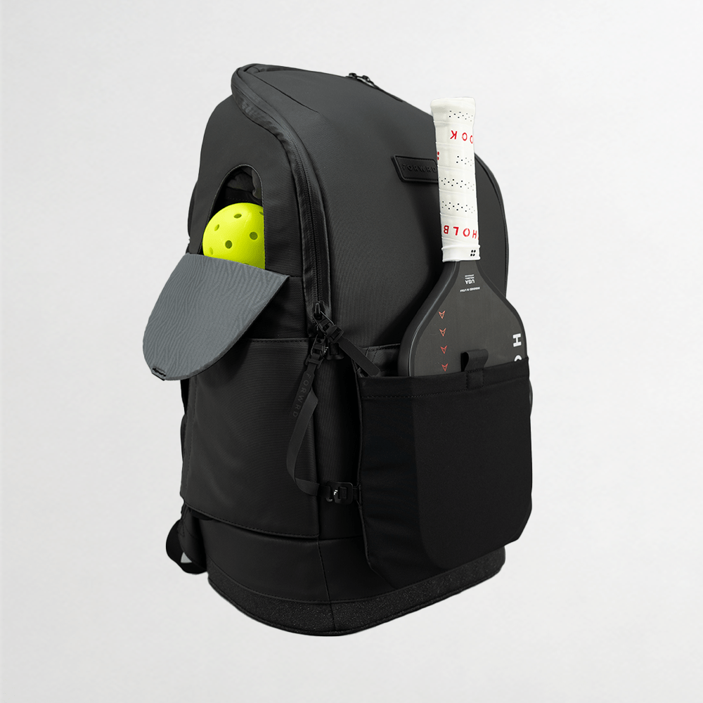 The FORWRD Court Caddy Front Paddle Sleeve, in black, holds a black pickleball paddle with white grip and red text. Its side pocket stores a yellow pickleball and a gray cap (brim out), all set against a plain light gray background.