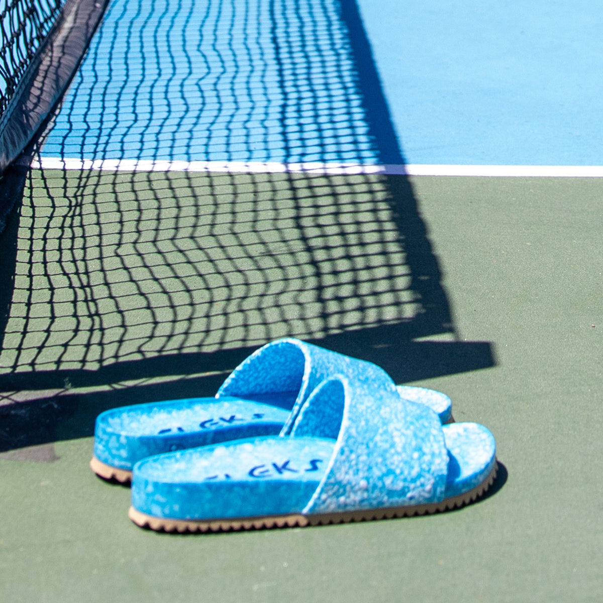 A pair of Blumaka Fleks® East Beach Slide blue slippers on a tennis court—ideal for athlete recovery and designed to ease soreness after intense matches.