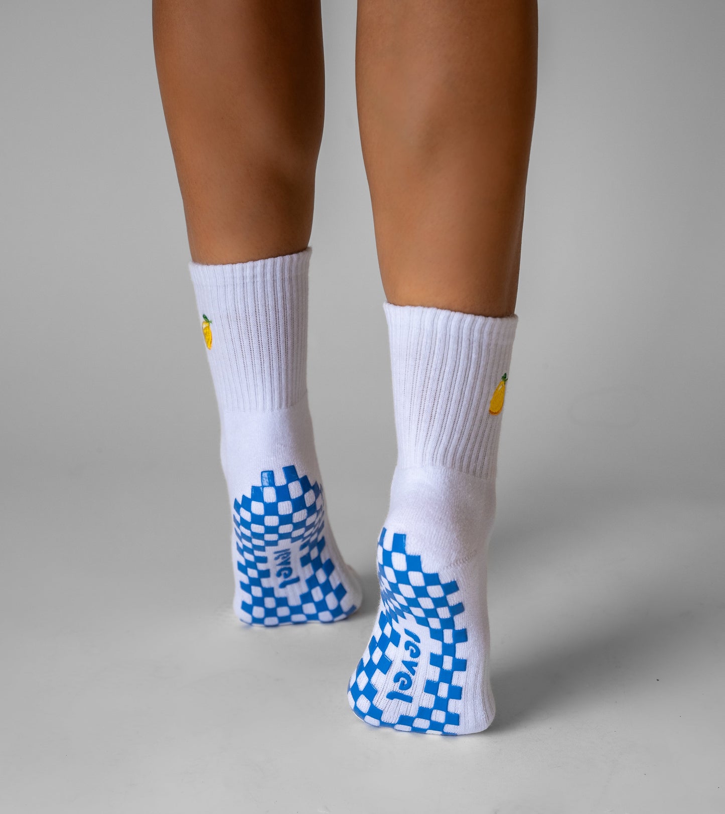 A person stands on their toes, displaying the backs of their legs and Revel's The Limoncello Grip Sock—white crew yoga socks with an embroidered lemon by the ankle, blue checkered Pilates grip soles, and "Play!" text on a plain light gray background.