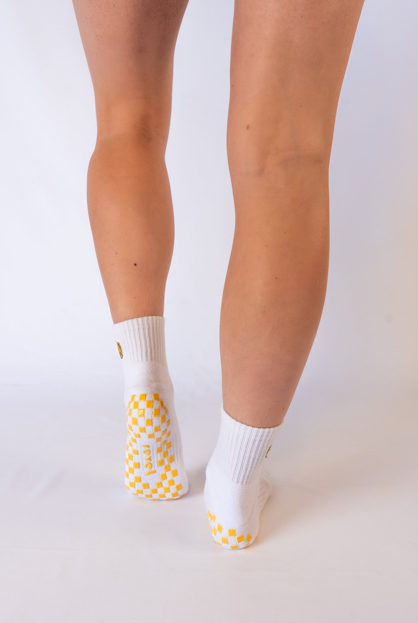 A person stands on tiptoe, viewed from behind, wearing Revel's The I'm Fine Grip Sock—white socks featuring yellow checkerboard grip soles and smiley faces on the back—against a seamless white background, showcasing only their lower legs and feet.