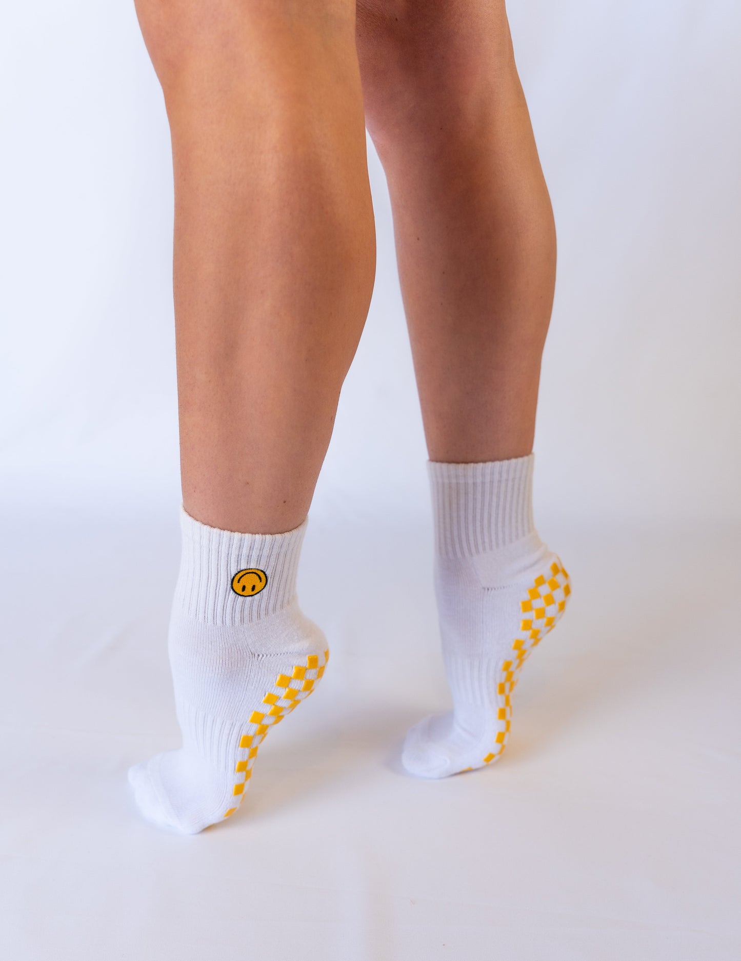 A person stands on tiptoes, seen from behind, wearing Revel's The I'm Fine Grip Sock. These white socks feature a yellow smiley face on the side and a yellow checkerboard pattern along the sole edges against a plain white background.