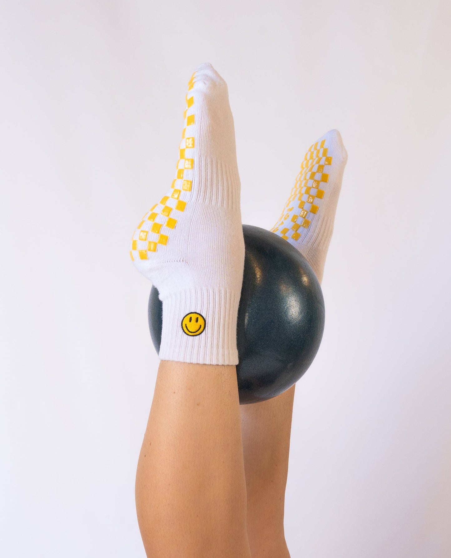 A person’s legs are raised, holding a dark green exercise ball, wearing Revel's The I'm Fine Grip Sock—white with a yellow checkerboard sole and a yellow smiley face on the ankle—set against a plain white background.