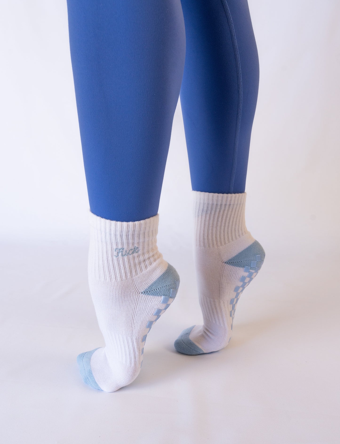 A person wears Revel's The Pardon My French Grip Sock—white with light blue non-slip grips—while standing on tiptoe, heels raised. Only the legs and textured sock soles are visible against a plain white background.