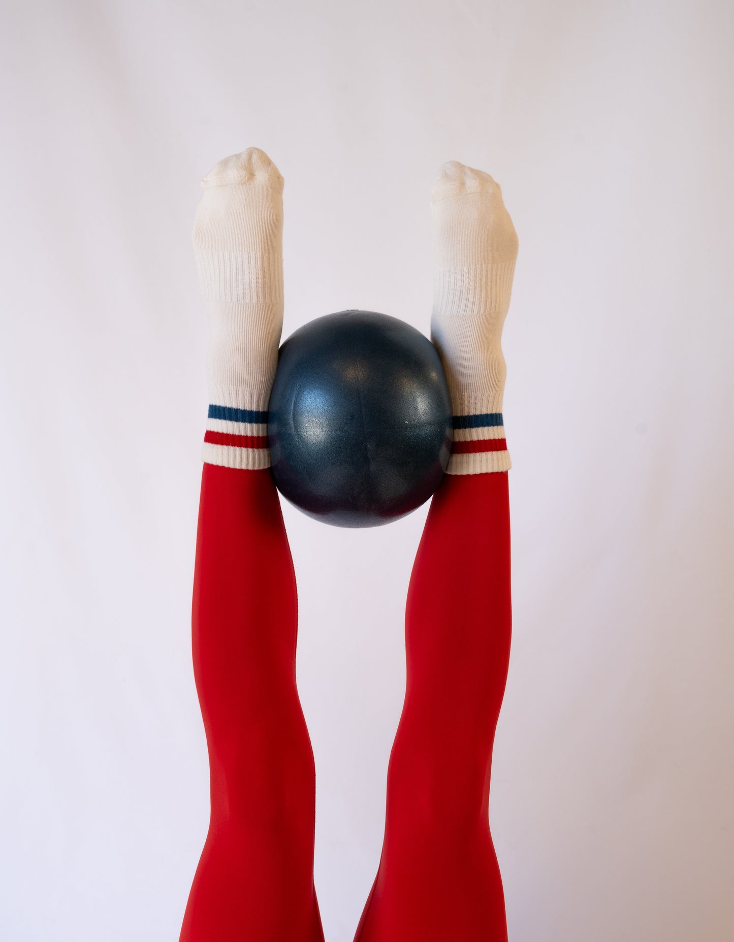 Two legs in bright red tights are raised vertically, with feet wearing Revel's The Miss Americana Grip Socks. The feet balance a dark blue ball between them, all set against a plain white background.