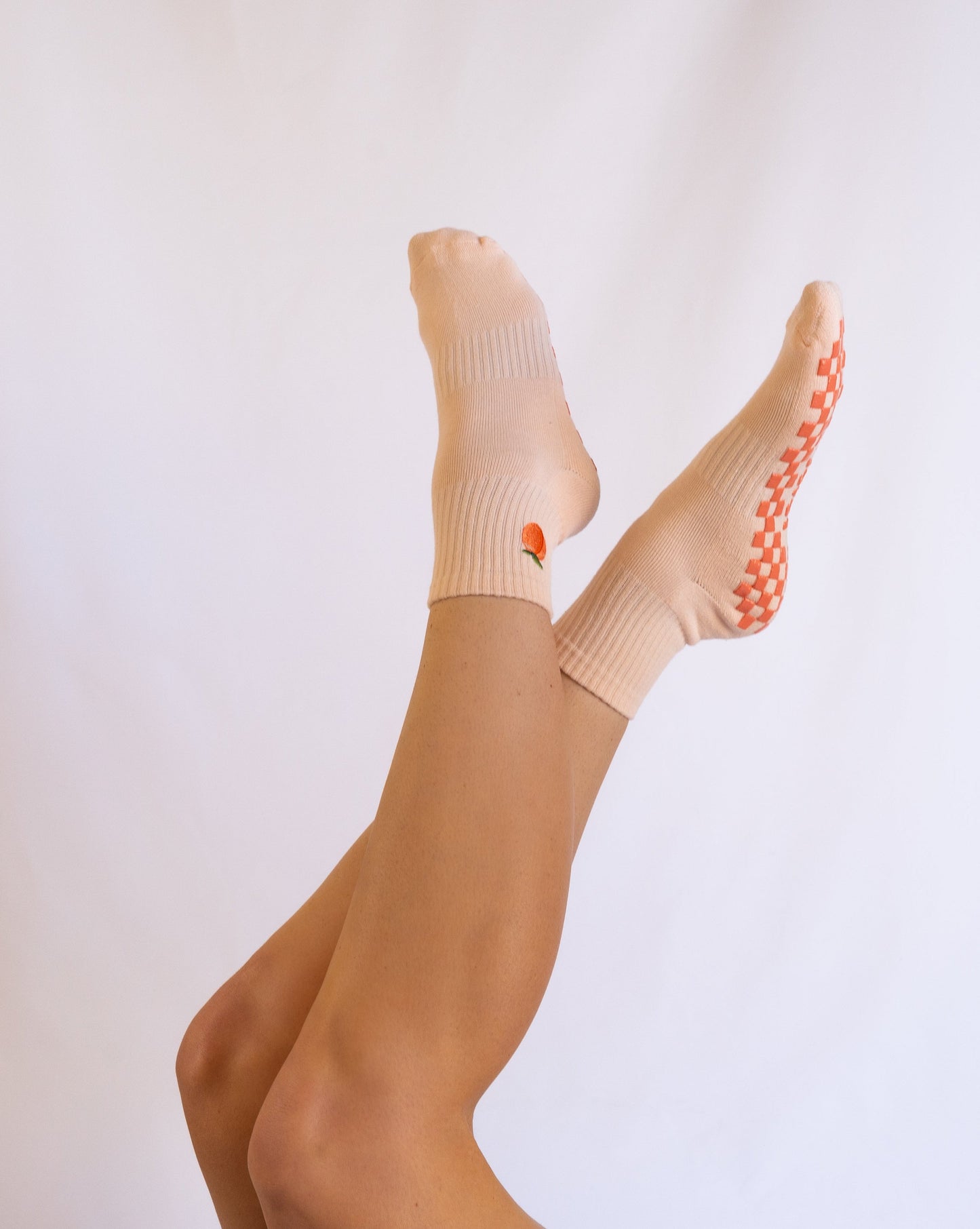 A person with light skin crosses their legs, showcasing Revel’s The Just Peachy Grip Sock in pale peach with orange grip dots and a small orange detail near the ankle against a white background.