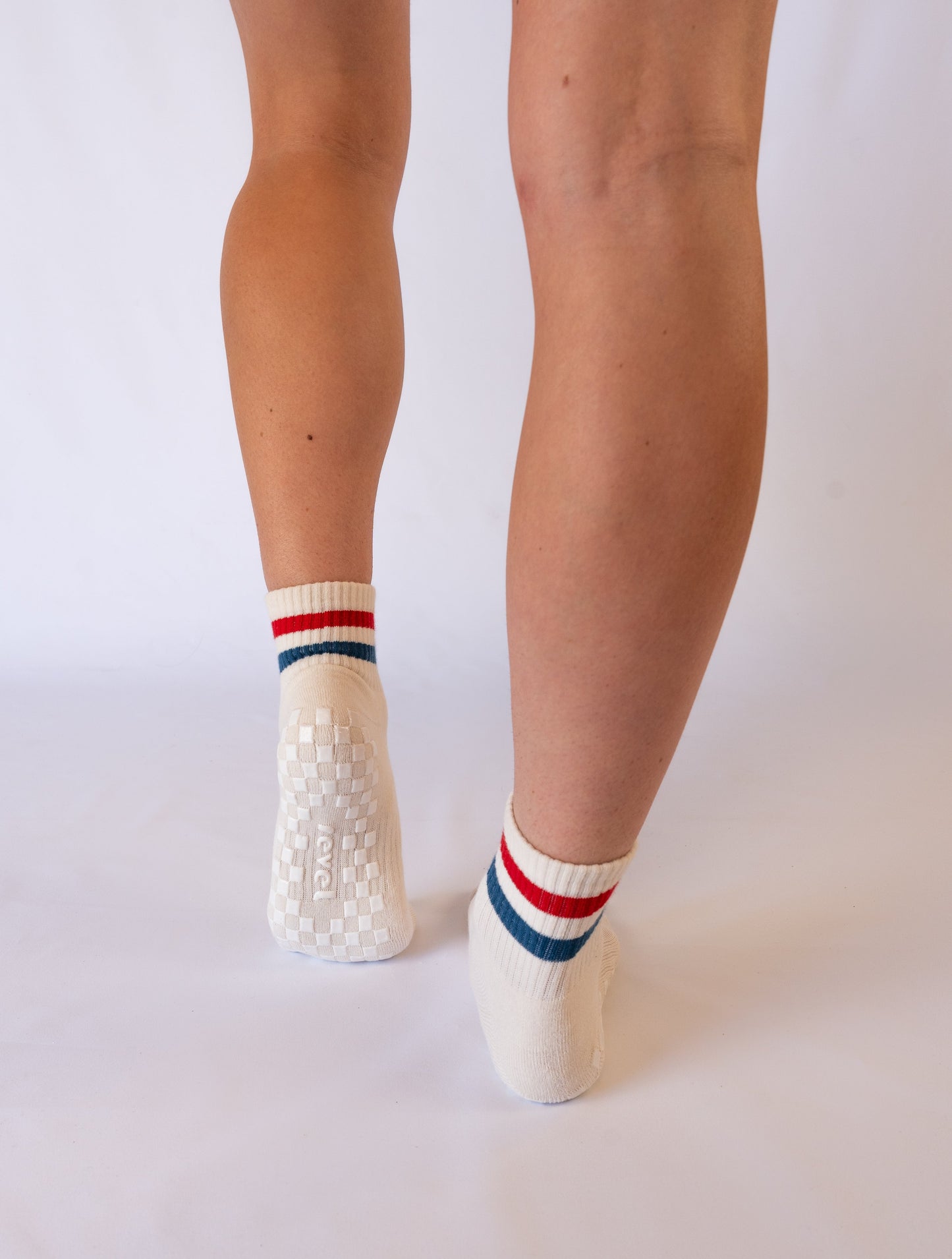 A person stands on tiptoe, viewed from behind, wearing Revel's The Miss Americana Grip Sock—white pilates socks with red and blue striped cuffs and a textured white non-slip grip sole. The plain white background shows just their lower legs and feet.