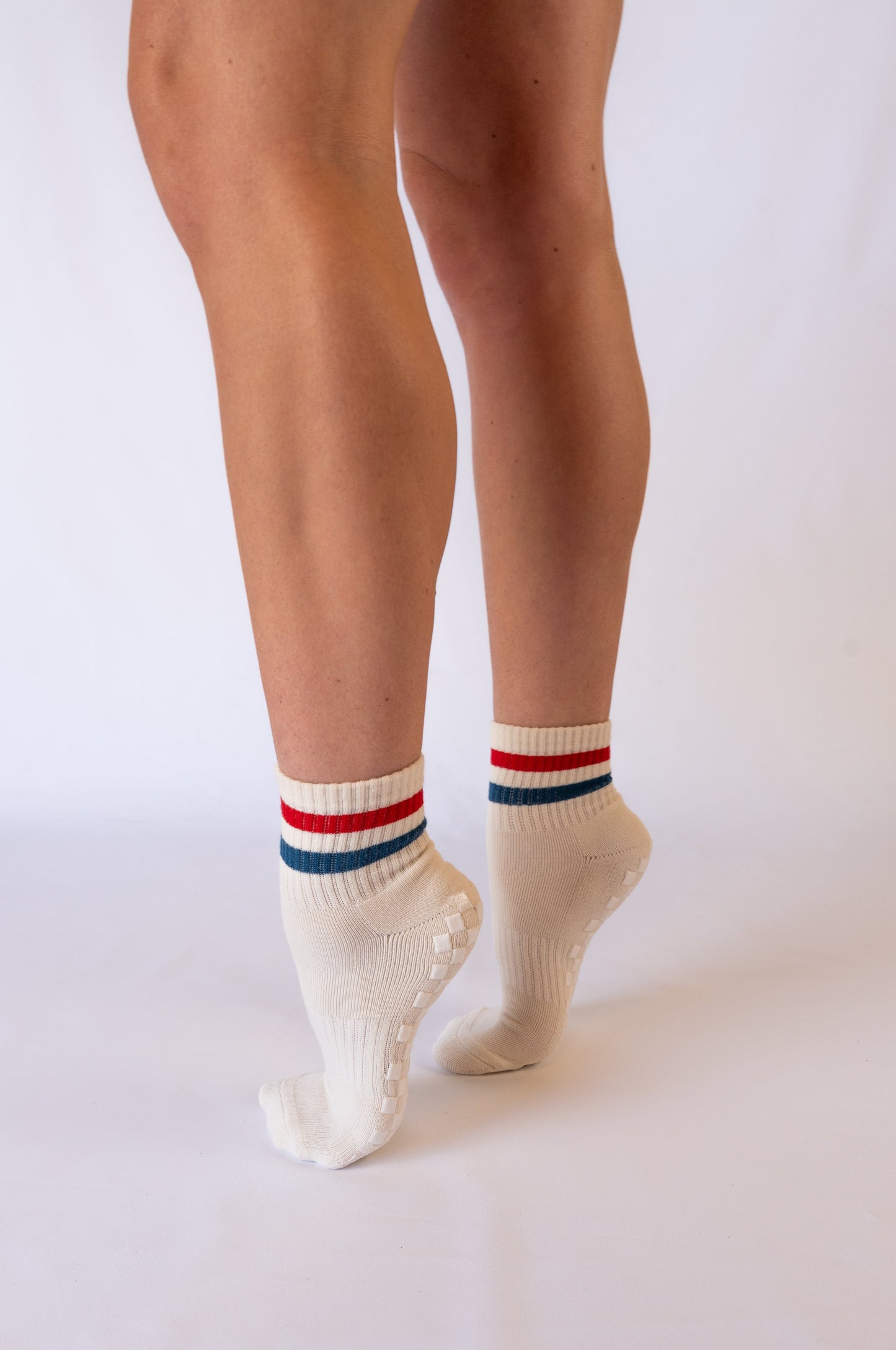 A person stands on tiptoe, revealing the backs of their bare legs in Revel's The Miss Americana Grip Sock—white ankle pilates socks with blue and red striped cuffs and small grippy dots on the soles—against a plain white background.