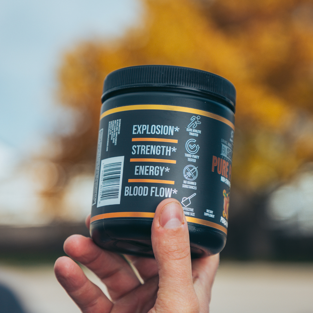 A hand holds a PURE POWER container by BLACKLABEL Supplements, with white label text highlighting explosion, energy, and blood flow benefits. This stimulant-free strength and endurance supplement has a ribbed black lid; autumn trees blur in the background.