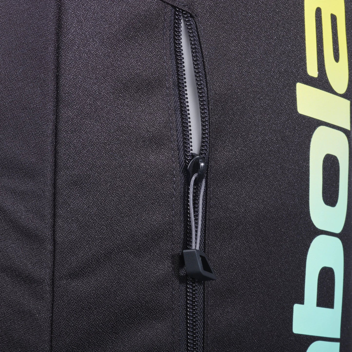 Close-up of the Babolat COURT Backpack LITE by Padel USA in black fabric, showing a vertical zipper with a black pull, and part of large yellow and green gradient text visible on the right.