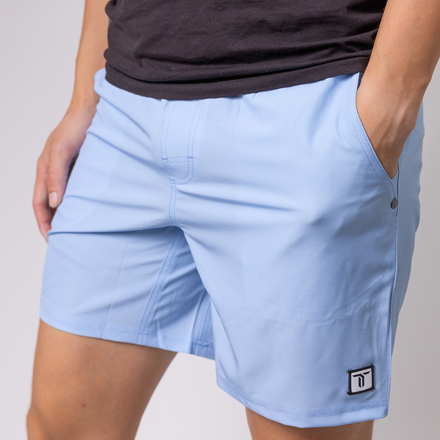 A person wearing light blue Thrive Pickleball Thrive Elite Shorts (7 in.) with a stitched black-and-white logo on the lower left leg stands with one hand in a pocket, paired with a black t-shirt. The background is plain and light-colored.