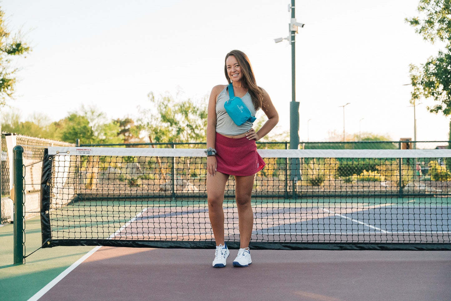 A woman smiles on an outdoor tennis court, wearing a tan tank top, maroon skirt, white sneakers, and the Thrive Pickleball Belt Bag crossbody. Trees and a bright sky complete the scene.