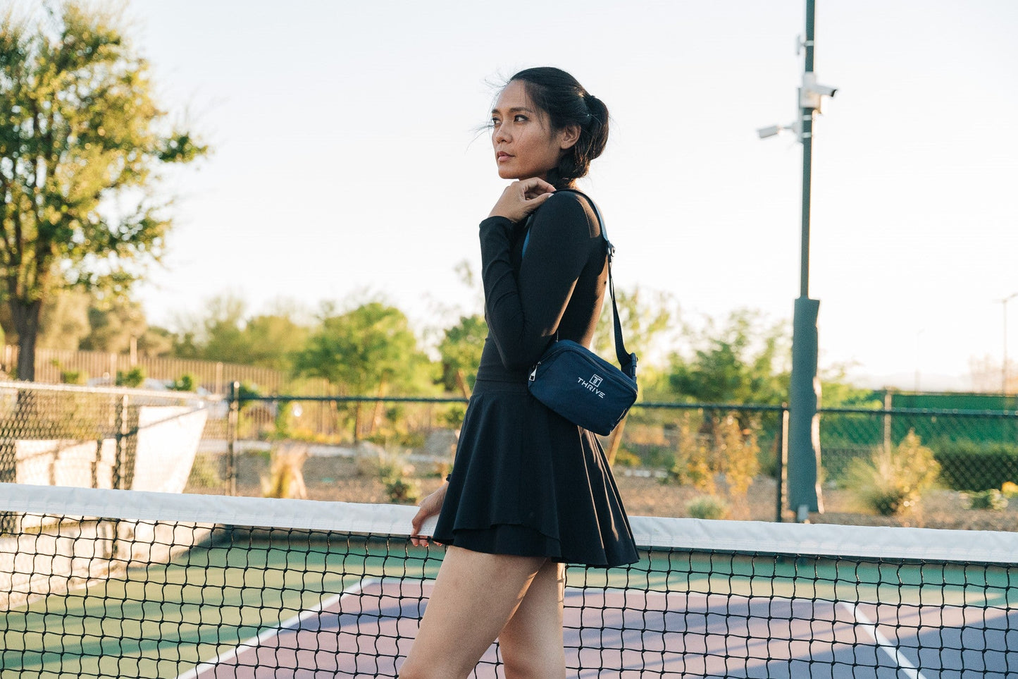 A woman in a black long-sleeve dress and the Thrive Pickleball Belt Bag stands by the tennis court net, gazing into the distance as sunlight filters through trees and a fence surrounding the court.