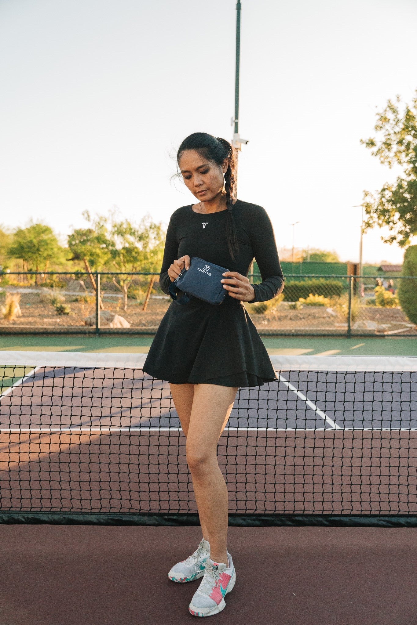 A woman stands on a tennis court in front of the net, wearing a black long-sleeve dress and colorful sneakers. She holds the Thrive Pickleball Belt Bag and is surrounded by trees under a clear sky.
