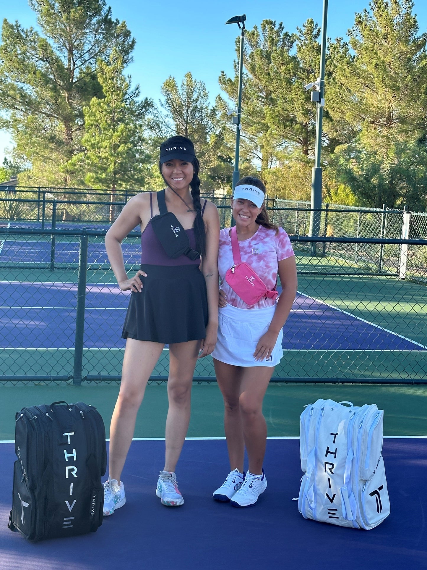 Two women pose on an outdoor tennis court, each wearing a Thrive Pickleball Belt Bag—one black, one pink—for organized travel. Both sport black THRIVE hats, with two white THRIVE tennis bags and sporty outfits completing the look.