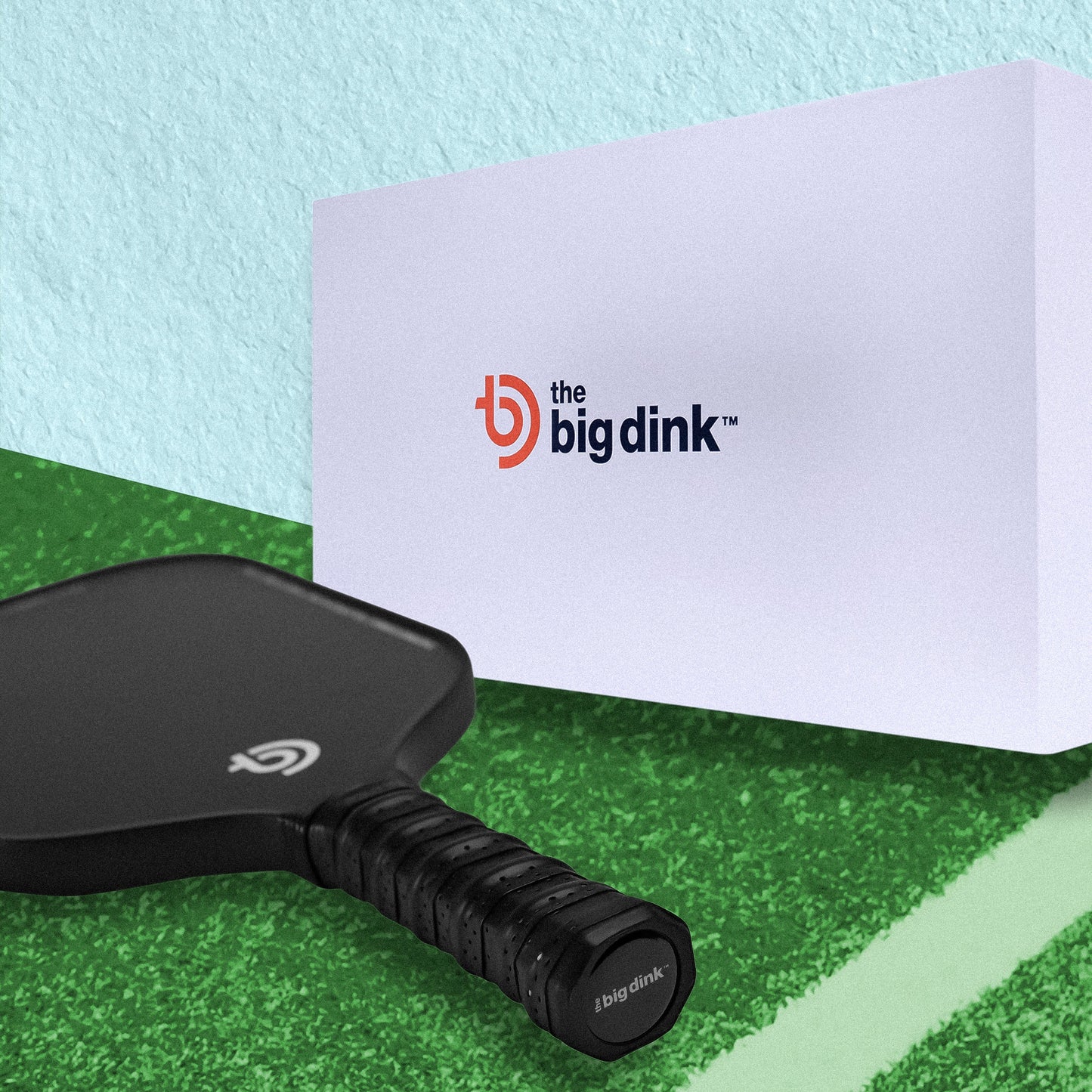 A black carbon fiber The Big Dink - Model2 Pickleball Paddle by The Big Dink rests on a green court. Behind it, a white box displays the brand’s blue and orange logo. Light blue and green walls create an ideal scene for spin lovers.