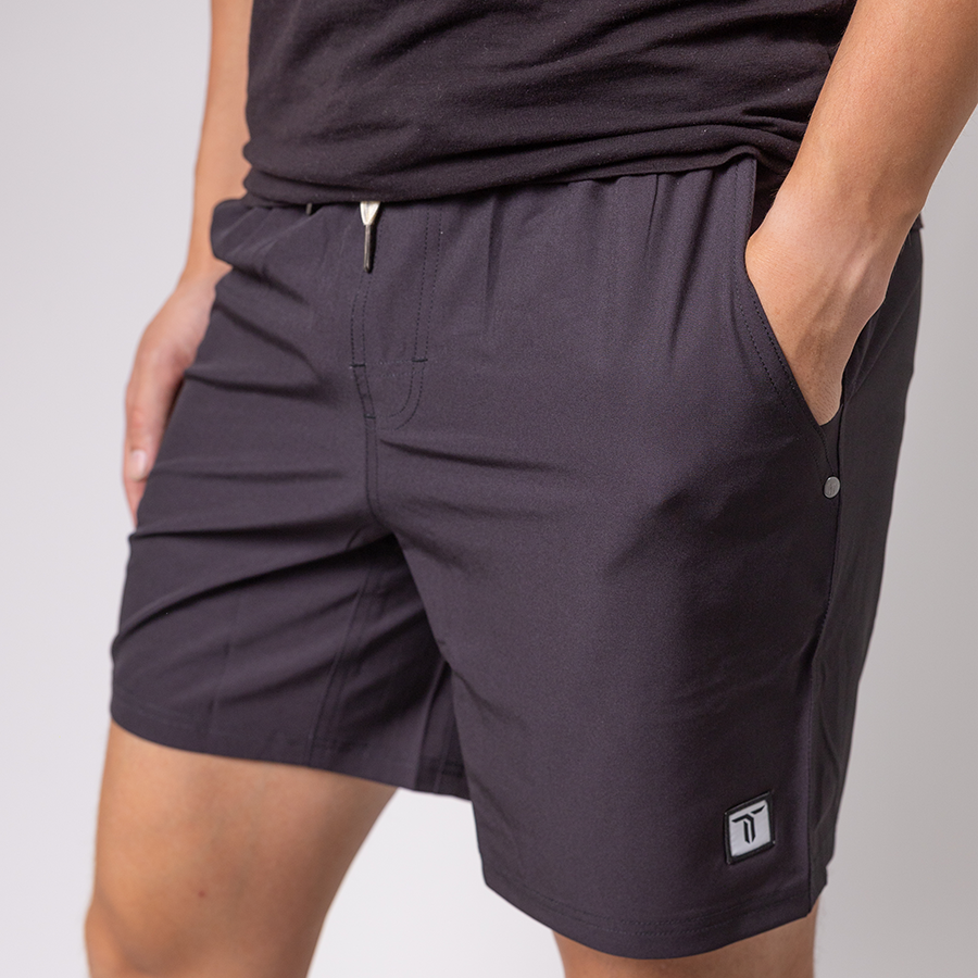 A person wears a black T-shirt and Thrive Elite Shorts (7 in.) by Thrive Pickleball, featuring lightweight stretch fabric, an elastic waistband with drawstring, and a small logo patch near the left hem. The background is plain and light-colored.