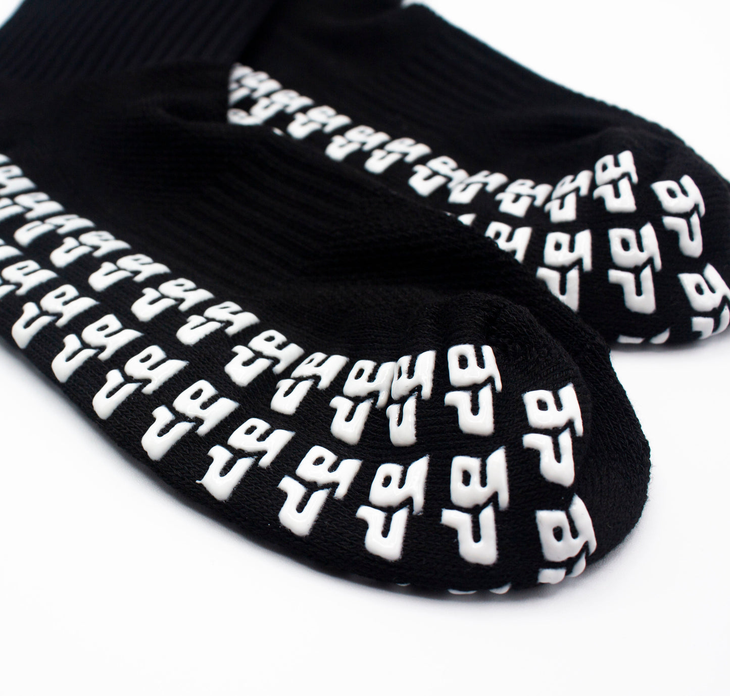 Close-up of Bodhi Performance’s GRIPtech™ Performance Socks in black, featuring white silicone geometric grip patterns on the soles for secure traction, displayed against a plain white background.