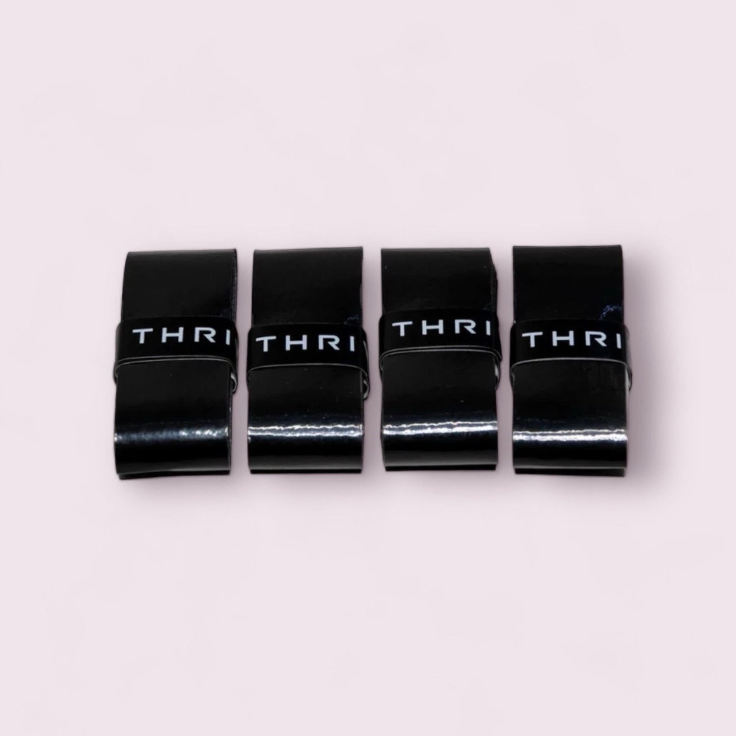 Four black rectangular Overgrips (Super Tacky) from Thrive Pickleball with glossy finishes are neatly aligned on a light pink background, each wrapped in a black band labeled “THRI,” suggesting their super tacky grip.