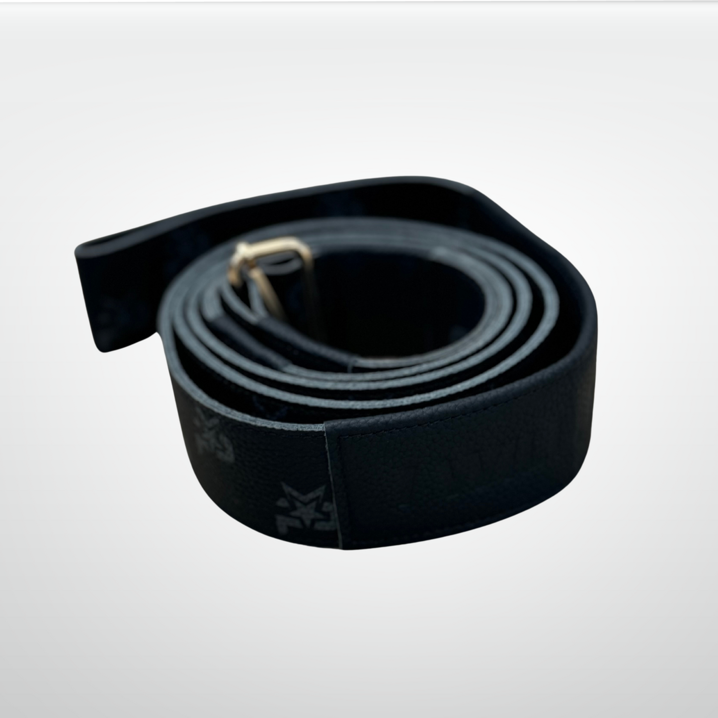 The iReliev I AM Leather Monogram Yoga Strap, featuring premium black textured leather and faint embossed branding, is coiled flat with a gold buckle visible against a smooth white-to-light-gray gradient background.