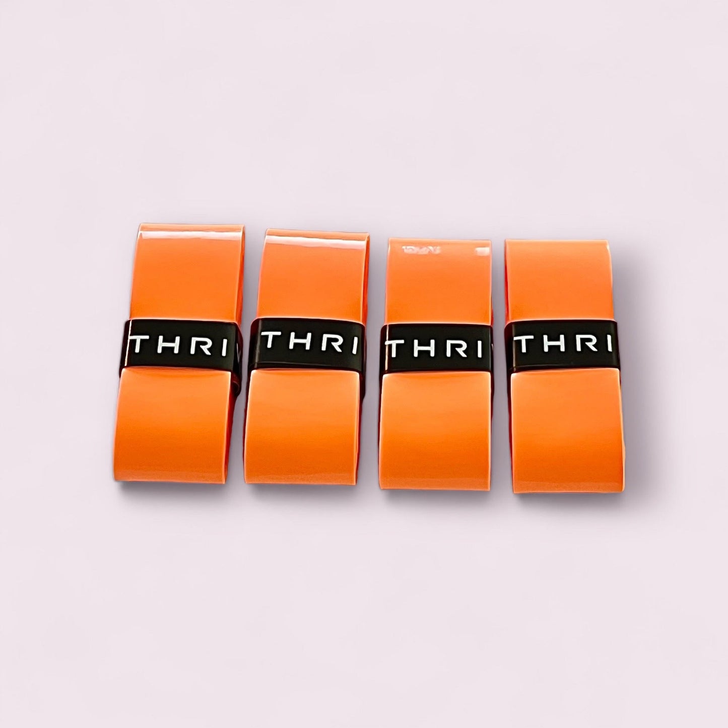 Four orange rectangular Overgrips (Super Tacky) by Thrive Pickleball, each with a glossy finish and a black band displaying “THRI,” are arranged in a row on a light pink background.