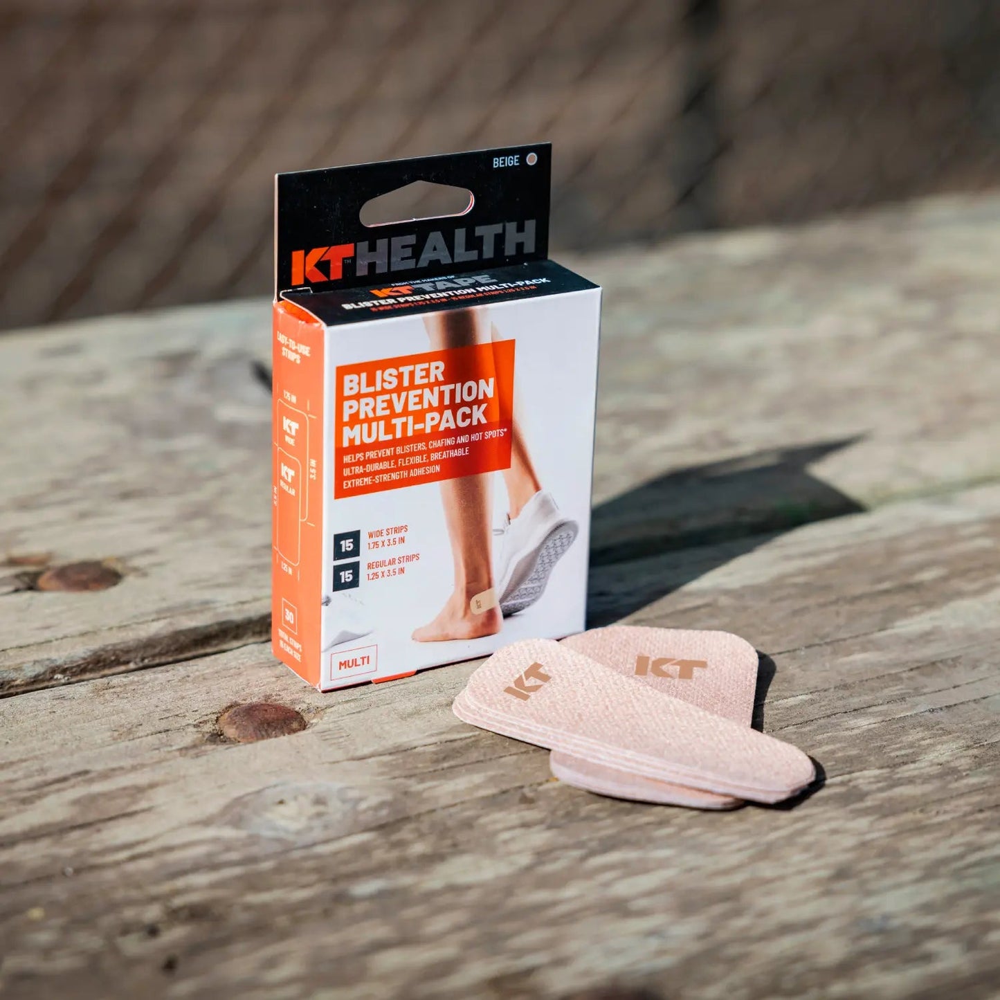 A box of KT Tape KT Health Blister Prevention Tape rests on a wooden surface outdoors, with several beige patches displayed in front. The pack contains 15 patches for heels, feet, and toes—ideal for athlete blister prevention.