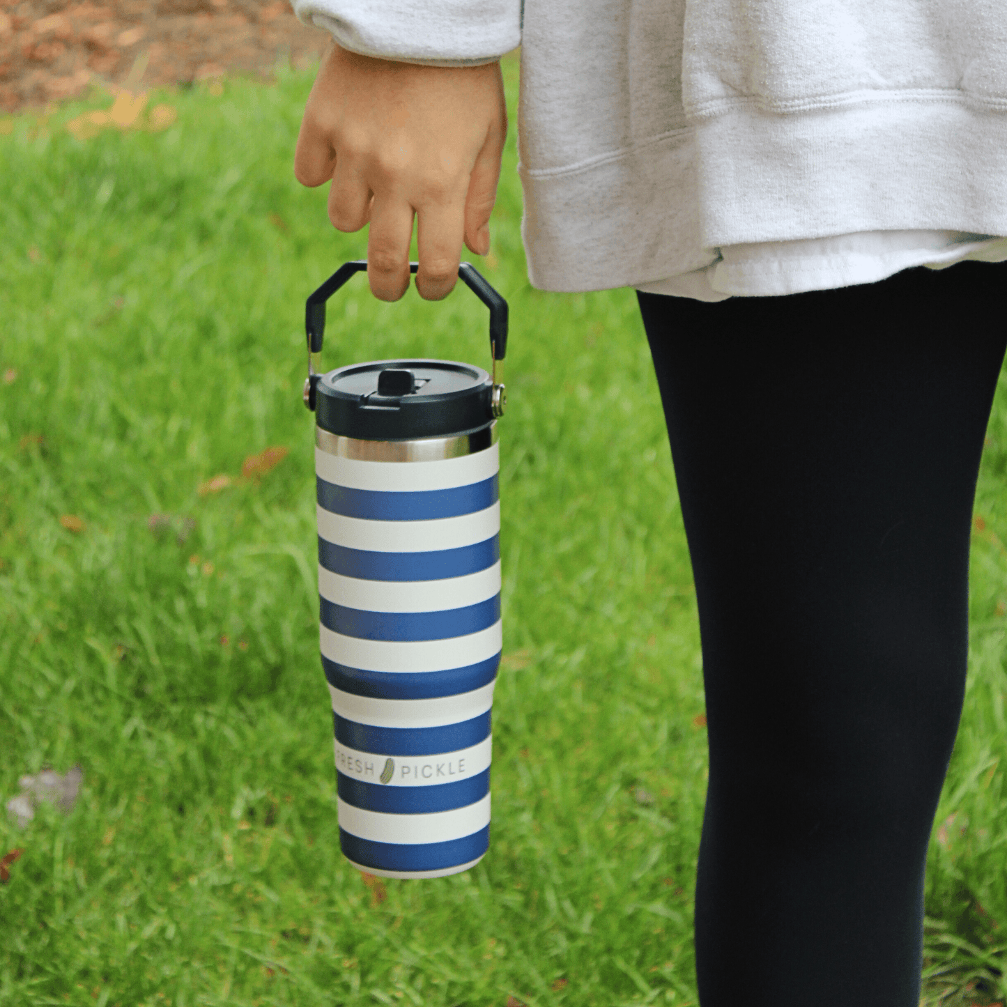 A person in a gray sweatshirt and black leggings holds the Fresh Pickle Tumbler by Fresh Pickle, a 30 oz blue-and-white striped water bottle with a flip-up straw, while standing outdoors on green grass; only their lower torso and hand are visible.