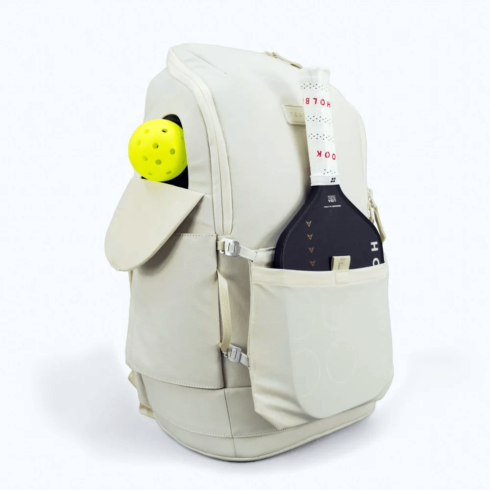 The FORWRD Court Caddy Pickleball Bag, in light beige and made from premium materials, features multiple pockets and holds a yellow pickleball plus a black paddle partly visible in an outer pocket, set against a plain light gray background.