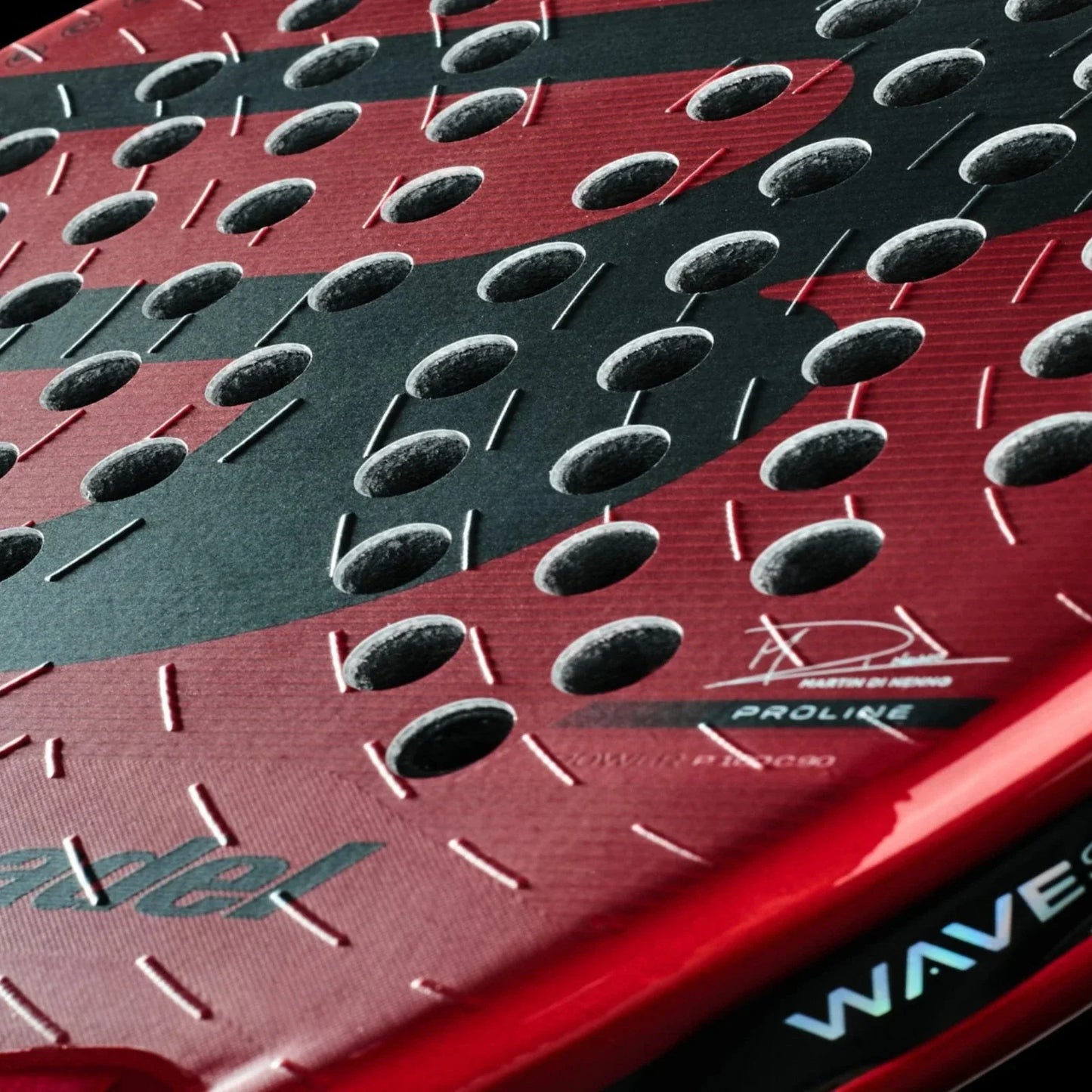 Close-up of the red Padel USA Bullpadel XPLO Comfort padel racket (2025), showing multiple circular holes, matte surface, embossed patterns for explosive power, and "WAVES" in blue and white on the edge.