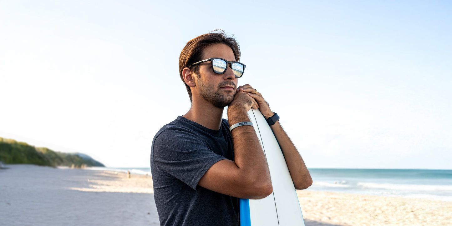 A man wearing Neven Eyewear Canova rectangle sunglasses and a dark t-shirt stands on a sandy beach holding a white surfboard, the sky reflected in his glasses as the ocean stretches beneath a clear blue sky.