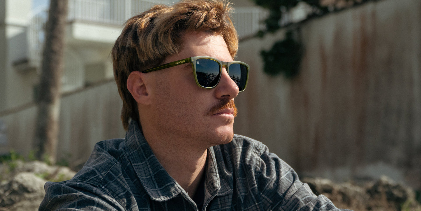 A man with light brown hair and a mustache wears Neven Eyewear's Fir green rectangle sunglasses and a plaid shirt, gazing thoughtfully into the distance with a blurred white building and greenery in the background.