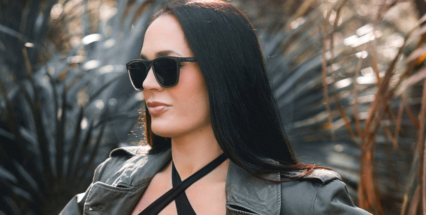 A woman with long straight black hair wears Neven Eyewear's Fir green rectangle sunglasses and a grey jacket, standing outdoors before blurred greenery as sunlight highlights her face and hair.