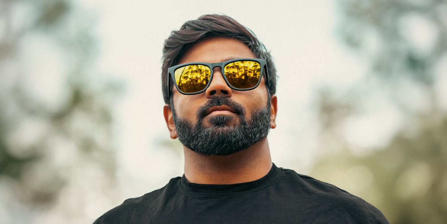 A bearded man with short dark hair wears Neven Eyewear’s Liquid Gold sunglasses with gold lenses and a black t-shirt, gazing confidently outdoors as the reflective lenses capture a vivid, leafy scene with green trees and sky behind him.