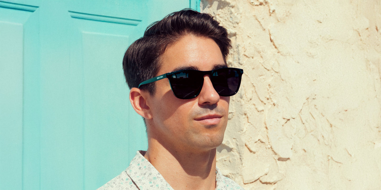 A man with short dark hair wears Neven Eyewear’s Nix black rectangle sunglasses with UV400 protection and a light-patterned shirt, standing in sunlight before a beige wall and turquoise door. His expression is neutral, evoking a summery vibe.