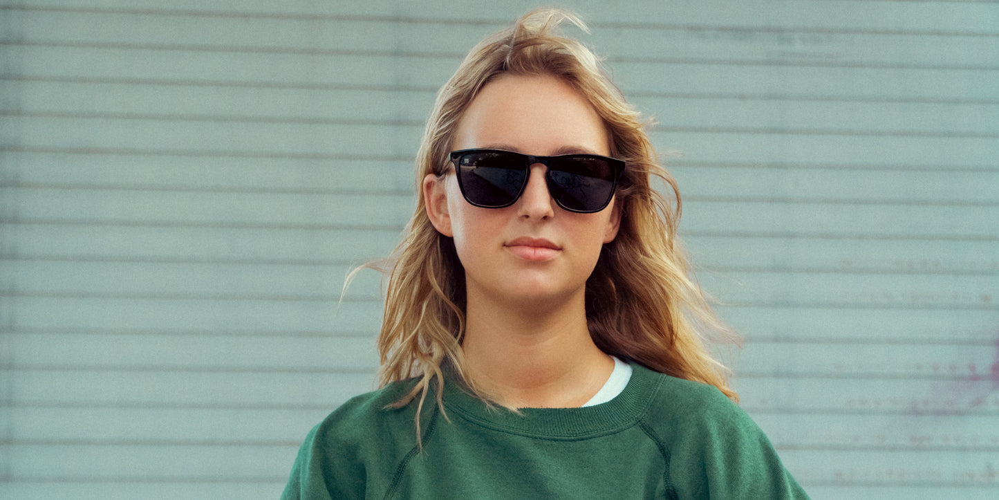 A young woman with long blonde hair faces forward, wearing Neven Eyewear's Nix black rectangle sunglasses with UV400 protection and a green sweatshirt, standing before a light-colored striped wall. Her hair appears slightly windswept.