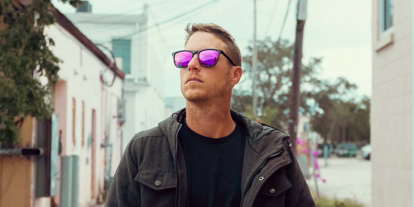 A man wearing Neven Eyewear's Violet sunglasses and a dark jacket walks through an alley lined with pastel buildings. He looks off to the side, with short hair, trees, power lines, and a cloudy sky in the background.