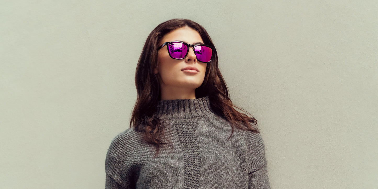 A woman with long dark hair wears a chunky gray sweater and Neven Eyewear's Violet polarized UV400 sunglasses. Against a plain light wall, she gazes upward, her loose hair falling over her shoulders and her expression neutral.