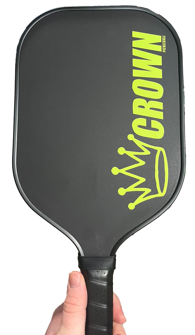 A hand holds the CROWN PICKLEBALL - Carbon Blade 13 (BLACK Paddle) by Crown Pickleball, featuring a black carbon fiber textured surface, bold yellow-green "CROWN" lettering, a crown graphic near the top right, and a black grip-wrapped handle.