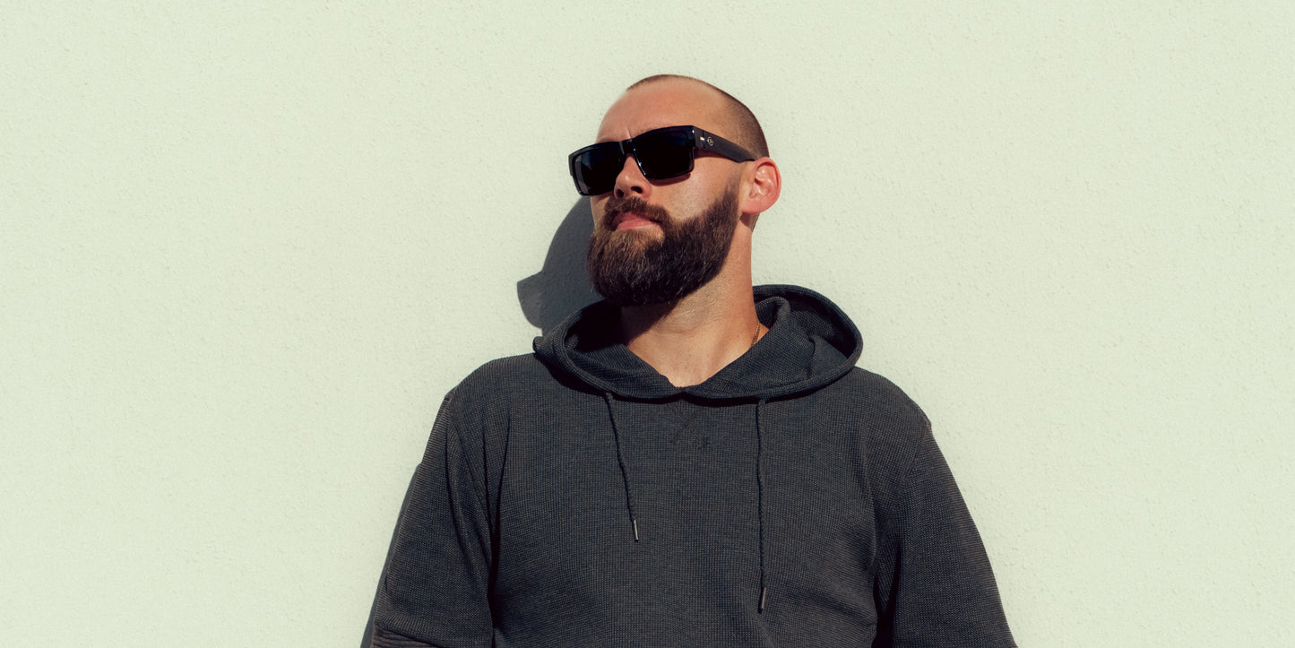 A man with a shaved head and full beard wears black Neven Eyewear Banshee sunglasses, part of the Catacombs collection. He stands against a plain light wall, facing slightly right with a neutral expression and a soft shadow behind him.