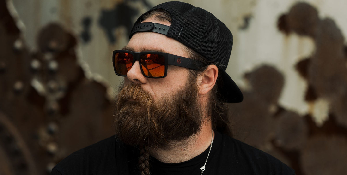 A man with a long brown beard and mustache wears a black cap backwards, a black shirt, and dark sunglasses with red lenses from the Neven Eyewear Havoc collection. His hair is braided, set against a background of weathered, rusty metal.