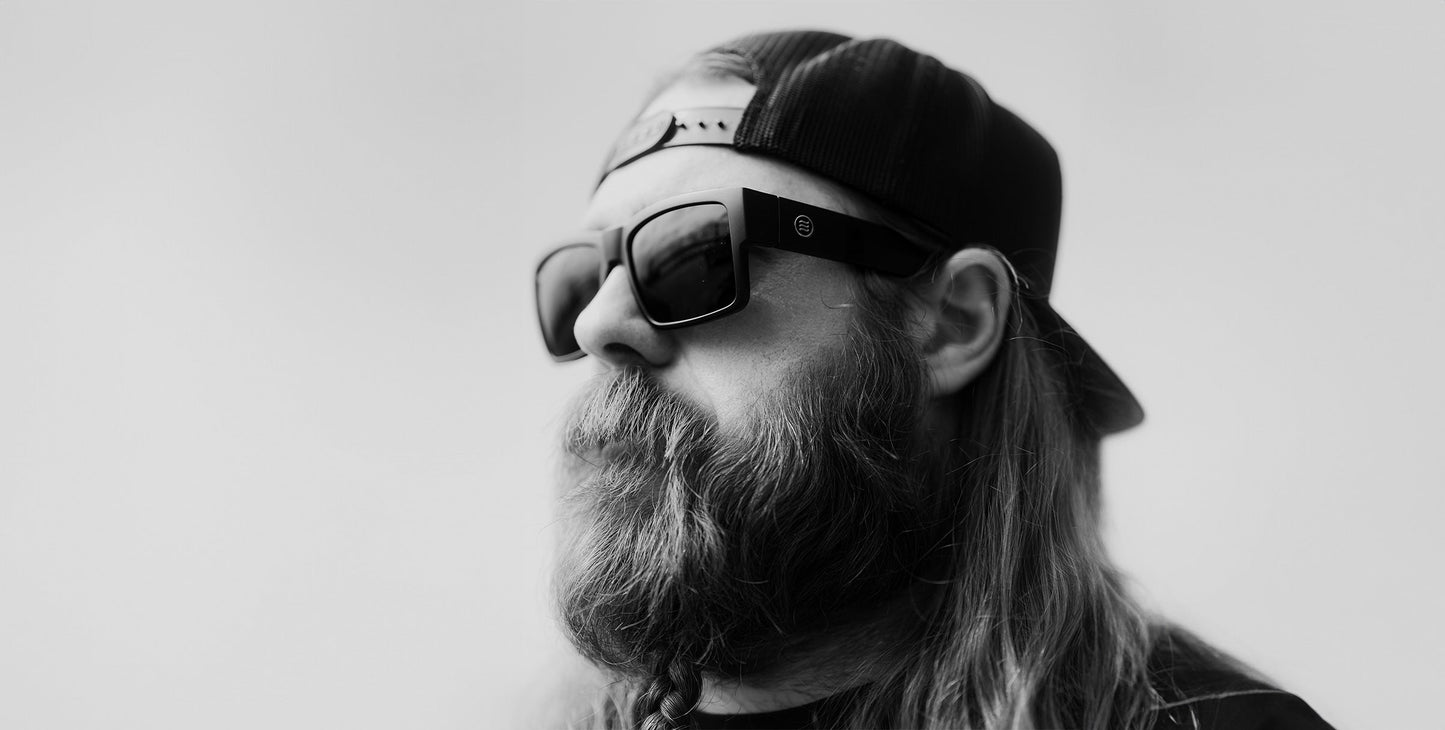 Black and white photo of a long-haired, bearded person in a backward cap, wearing bold Relic sunglasses from Neven Eyewear. They gaze to the left against a plain blurred background.