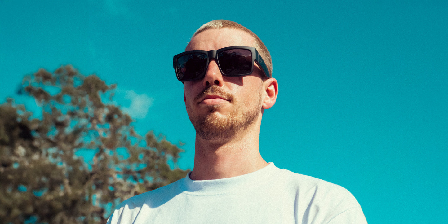 A man with a short beard and light hair, wearing Neven Eyewear Relic sunglasses and a white t-shirt, stands outdoors under a bright blue sky. Trees blur in the background as sunlight casts shadows on his face.