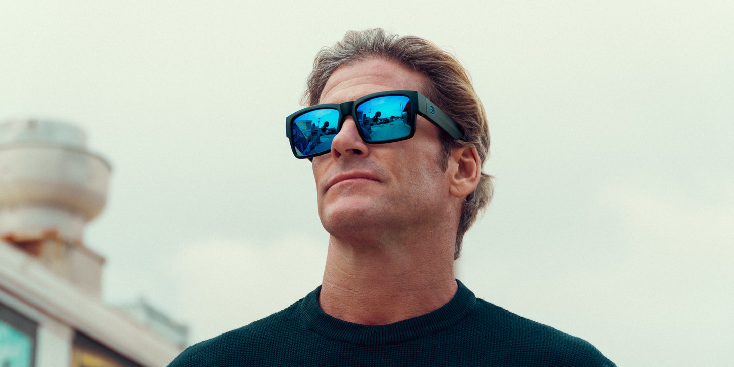 A man with light brown hair wears Neven Eyewear Scythe sunglasses with mirrored blue lenses and a dark green sweater, gazing upward with a neutral expression. An overcast sky and part of a building appear in the background on the left.