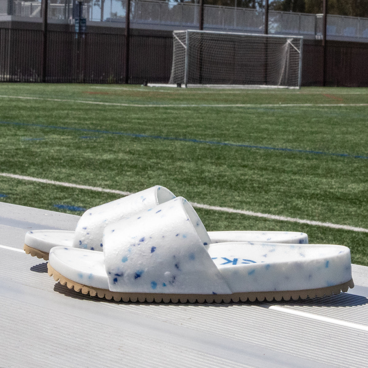 A pair of Blumaka Fleks® East Beach Slides—white with blue speckles and textured soles for alignment—rests on metal bleachers next to an empty artificial turf soccer field, with a goalpost and fence in the background.