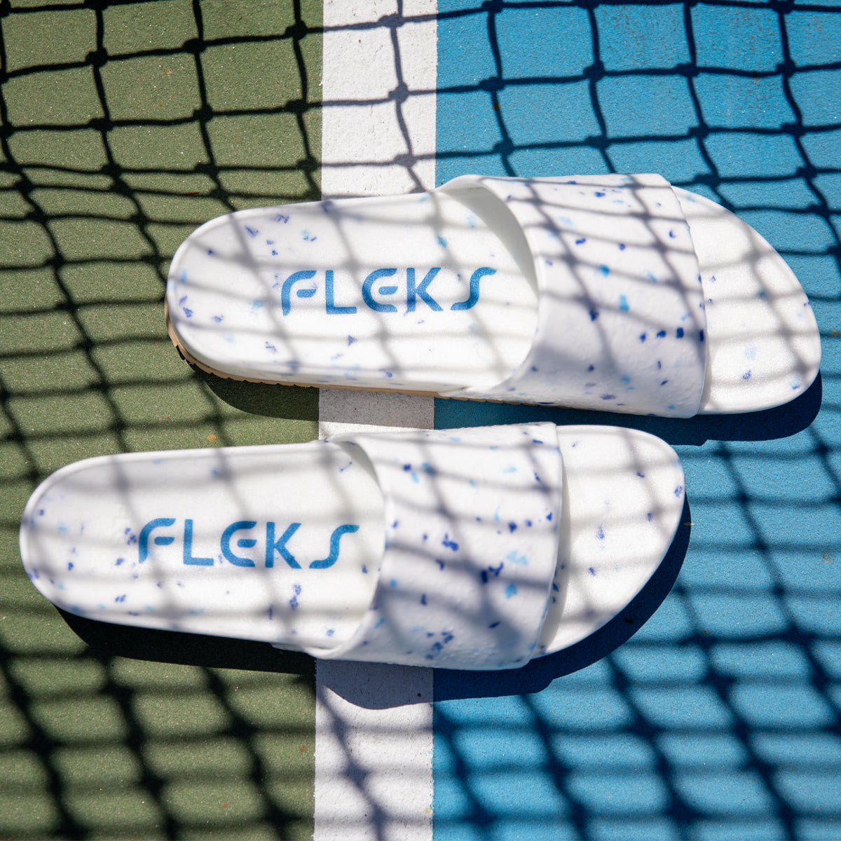The Blumaka Fleks® East Beach Slide, white with blue speckles and "FLEKS" in blue, rests on a tennis court beneath net shadows—designed for alignment and athlete recovery.