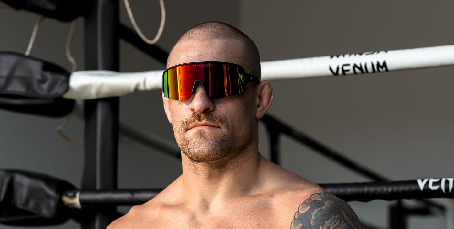 A muscular, shirtless man with a shaved head and mustache stands in a boxing ring wearing Neven Eyewear Cadence running sunglasses with polarized UV400 protection. Boxing ropes and a VENUM corner post can be seen in the bright background.